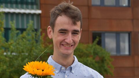 Absolvent vor dem Dienstgebäude mit Sonnenblume und Zeugnis in der Hand