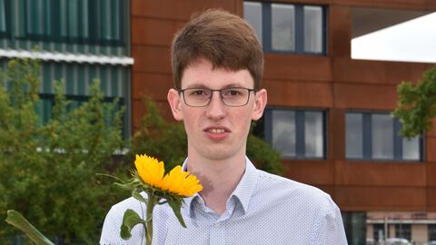 Absolvent vor dem Dienstgebäude mit Sonnenblume und Zeugnis in der Hand