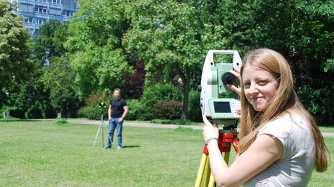 Geomatikerin steht mit Tachymeter auf Wiese