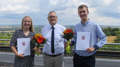 Zwei Absolventen mit Zeugnissen und Blumen, Amtsleitung in der Mitte
