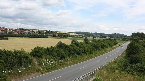 Eine Straße mit einem Auto führt an einem Feld vorbei, im Hintergrund sind Häuser