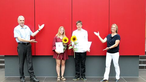 Amtsleitung und Ausbilderin mit den zwei Absolventen vor einer roten Wand