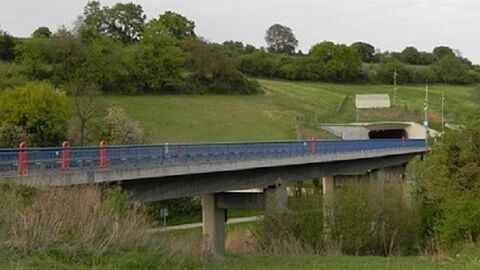 Eine Brücke mit blauem Geländer inmitten grüner Landschaft führt in einen Tunnel, 