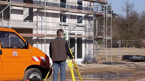 Gebäudeeinmessung Rückenansicht eines Mannes an einem Messinstrument, im Hintergrund ein im Bau befindliches Haus, links daneben ein orangefarbener Messbus
