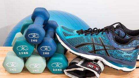 Sport und Gesundheit 6 Hanteln aufeinandergelegt, daneben ein Paar Turnschuhe, im Hintergrund ein Sitzball