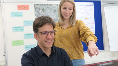 Zwei Personen vor einem Laptop, eine sitzt vor dem Laptop, die stehende Person deutet auf den Monitor