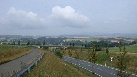 Eine Straße verläuft in einem Linksbogen zwischen Feldern unter einer Brücke hindurch. Links etwas oberhalb verläuft ein befestigter Weg