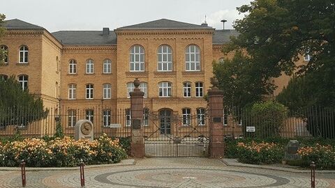 Gros helles Backsteingebäude, davor eine Zufahrt mit Gittertor