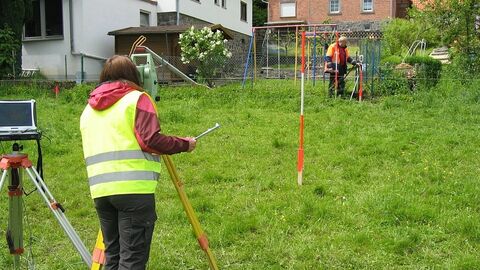 Zwei Personen mit Messinstrumenten auf einer Wiese an einem Haus