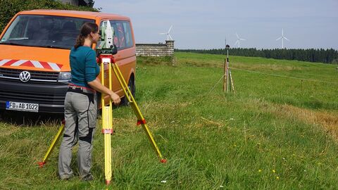 Person mit Messinstrument auf Wiese, im Hintergrund ein orangefarbener Messbus