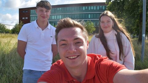 3 junge Menschen im Freien blicken in die Kamera, einer im Vordergrund, zwei seitlich dahinter