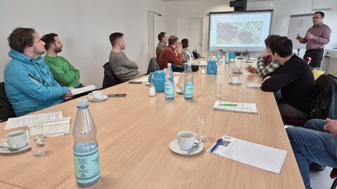 Mehrere Personen sitzen um einen Tisch, eine Person steht neben einem Whiteboard